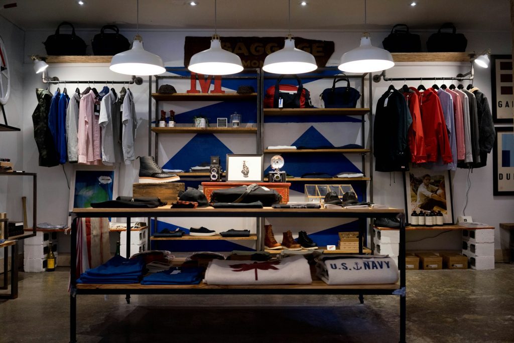 Interior de uma pequena loja de roupas com araras, prateleiras e luminárias