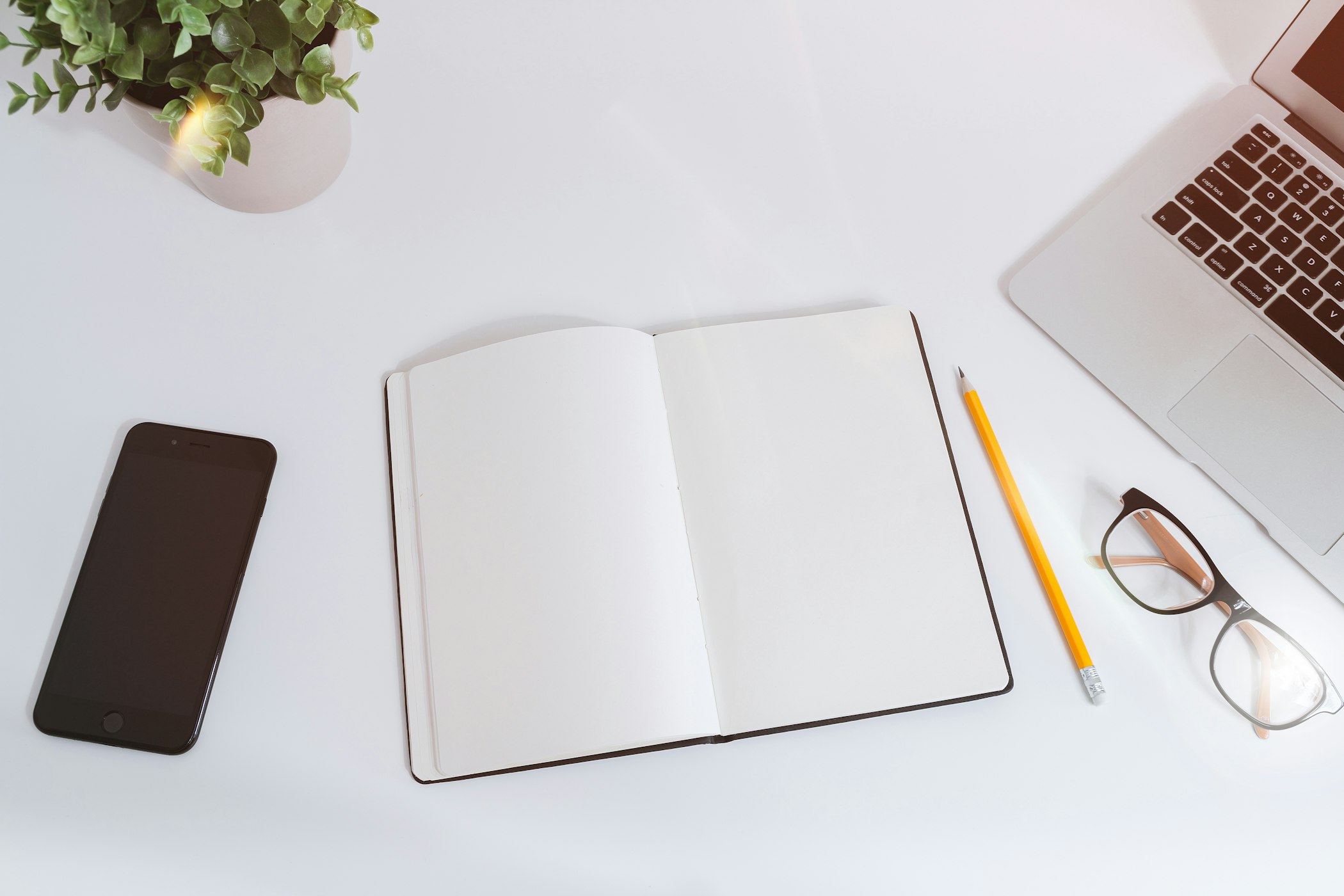 Caderno aberto ao lado de notebook, lápis e óculos sobre uma mesa branca